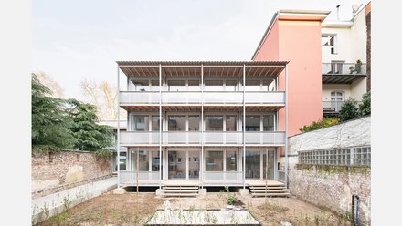 Rückfassade eines dreigeschossigen Wohnbaus mit umlaufenden Laubengängen aus Metall; kleiner Hof mit junger Bepflanzung, rosa Nachbarhaus.