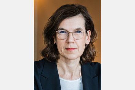 Porträt einer Frau mit Brille, schulterlangen dunklen Haaren und dunklem Blazer vor warmem Hintergrund.