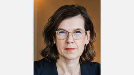 Porträt einer Frau mit Brille, schulterlangen dunklen Haaren und dunklem Blazer vor warmem Hintergrund.