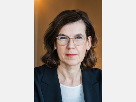 Porträt einer Frau mit Brille, schulterlangen dunklen Haaren und dunklem Blazer vor warmem Hintergrund.