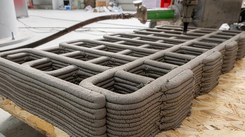Teilweise fertiggestellte 3D-gedruckte Lehmstruktur mit abgerundeten Ecken und innerem Raster, gefertigt durch robotische Extrusion.“