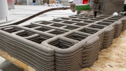 Teilweise fertiggestellte 3D-gedruckte Lehmstruktur mit abgerundeten Ecken und innerem Raster, gefertigt durch robotische Extrusion.“