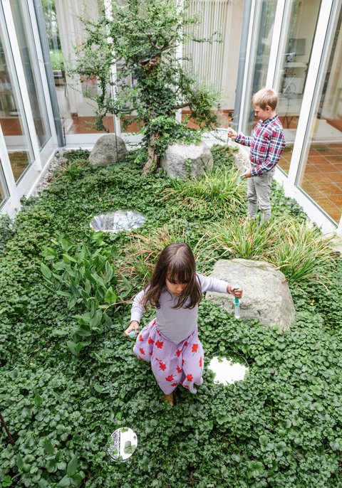 „Zwei Kinder spielen in einem hellen, verglasten Innenhof, der mit üppigen grünen Pflanzen, kleinen Felsen und runden Trittsteinen wie ein natürlicher Garten angelegt ist.“