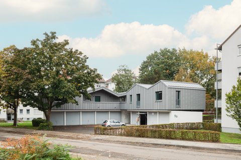 Aufgestocktes Wohngebäude mit grauer Metallfassade über Garagen, eingebettet in grüne Wohnsiedlung.