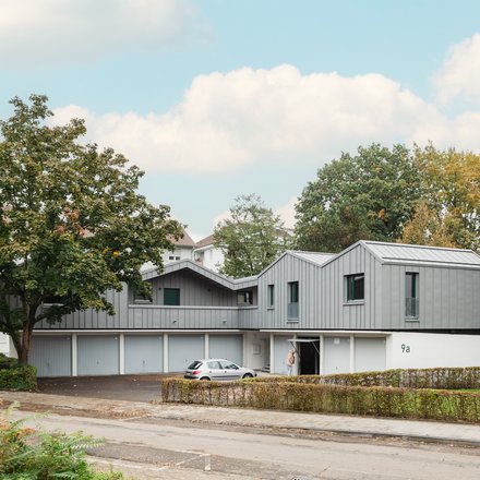 Aufgestocktes Wohngebäude mit grauer Metallfassade über Garagen, eingebettet in grüne Wohnsiedlung.