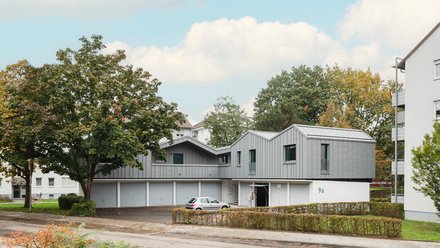 Aufgestocktes Wohngebäude mit grauer Metallfassade über Garagen, eingebettet in grüne Wohnsiedlung.