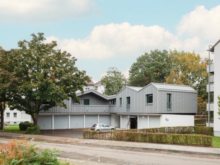 Aufgestocktes Wohngebäude mit grauer Metallfassade über Garagen, eingebettet in grüne Wohnsiedlung.