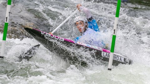 Person im Kajak fährt durch Wildwasser zwischen Slalomstangen, während Wasser über Boot und Paddel spritzt.