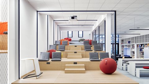 Moderner, heller Mehrzweckraum mit gestuften Sitzflächen, grauen und roten Kissen, rotem Gymnastikball und Bücherregalen im Hintergrund