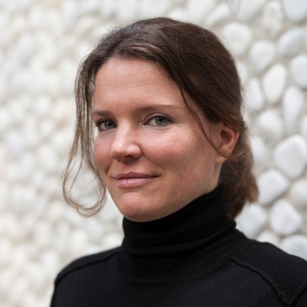 Porträt einer Frau mit schulterlangen braunen Haaren, die über die Schulter schaut. Sie trägt einen schwarzen Rollkragenpullover. Der Hintergrund zeigt weiße und hellgraue Steinelemente, die leicht unscharf sind.