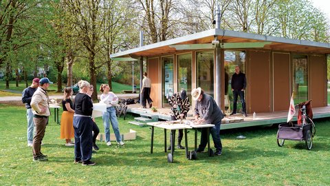"Mehrere Personen stehen an einem sonnigen Tag vor einem Holzpavillon mit Glasfronten zusammen. Einige betrachten in einer Gruppe Baupläne, andere arbeiten an einem Tisch mit Werkzeug. Ringsum stehen große Bäume."