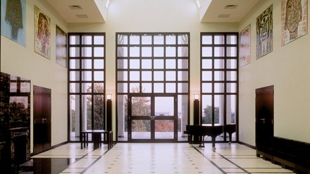 "Moderner Ausstellungsraum mit hochgewölbter Decke und rautenförmigen Oberlichtern in zwei Reihen. Creme-farbene Wände, große dreiteilige Fensterfront mit Ausblick auf herbstliche Vegetation. Vier dekorative Kunstobjekte an den Seitenwänden. Glatte Bodenfliesen mit Mosaikmuster. Zwei schwarze Stühle im Raum."