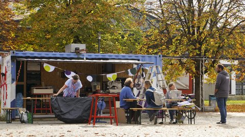 "Ein geöffneter blauer Schiffscontainer dient auf einem Platz als Werkstatt. Eine Wimpelkette hängt an der Vorderseite. Davor sitzen und stehen Personen an einem Tisch und unterhalten sich. Die Bäume ringsum tragen buntes Herbstlaub."
