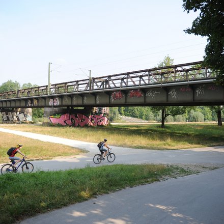 Ein Foto zeigt eine Brücke über eine Grünfläche mit einem Rad- und Fußweg, auf dem mehrere Menschen Fahrrad fahren, während darüber eine Bahntrasse verläuft; das Bild veranschaulicht die gleichzeitige Nutzung von Infrastruktur für Verkehr, Freizeit und Landschaft und steht für die Verbindung von Mobilität und öffentlichem Raum.