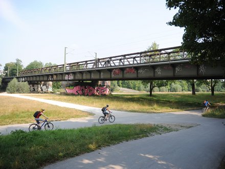 Ein Foto zeigt eine Brücke über eine Grünfläche mit einem Rad- und Fußweg, auf dem mehrere Menschen Fahrrad fahren, während darüber eine Bahntrasse verläuft; das Bild veranschaulicht die gleichzeitige Nutzung von Infrastruktur für Verkehr, Freizeit und Landschaft und steht für die Verbindung von Mobilität und öffentlichem Raum.