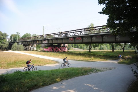 Ein Foto zeigt eine Brücke über eine Grünfläche mit einem Rad- und Fußweg, auf dem mehrere Menschen Fahrrad fahren, während darüber eine Bahntrasse verläuft; das Bild veranschaulicht die gleichzeitige Nutzung von Infrastruktur für Verkehr, Freizeit und Landschaft und steht für die Verbindung von Mobilität und öffentlichem Raum.