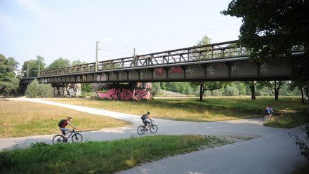 Ein Foto zeigt eine Brücke über eine Grünfläche mit einem Rad- und Fußweg, auf dem mehrere Menschen Fahrrad fahren, während darüber eine Bahntrasse verläuft; das Bild veranschaulicht die gleichzeitige Nutzung von Infrastruktur für Verkehr, Freizeit und Landschaft und steht für die Verbindung von Mobilität und öffentlichem Raum.
