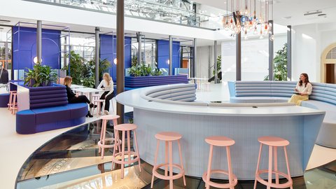 Begegnungszone mit Loungemöbeln: Menschen sitzen und sprechen an runden Sitzinseln, umgeben von Pflanzen und Glasdach.