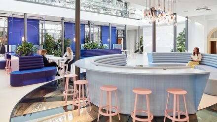 Begegnungszone mit Loungemöbeln: Menschen sitzen und sprechen an runden Sitzinseln, umgeben von Pflanzen und Glasdach.