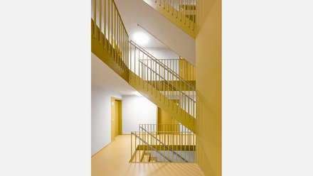 Modernes Treppenhaus mit ochkergelben Wänden und Geländern. Mehrere Treppenlauf mit metallischen Handläufen führen nach oben. Links eine gelbe Tür im Flur. Die Decke ist weiß mit integrierten runden Leuchten. Der Boden ist hellgelb. Das Treppenhaus ist hell und minimalistisch gestaltet.