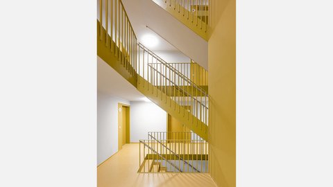 Modernes Treppenhaus mit ochkergelben Wänden und Geländern. Mehrere Treppenlauf mit metallischen Handläufen führen nach oben. Links eine gelbe Tür im Flur. Die Decke ist weiß mit integrierten runden Leuchten. Der Boden ist hellgelb. Das Treppenhaus ist hell und minimalistisch gestaltet.