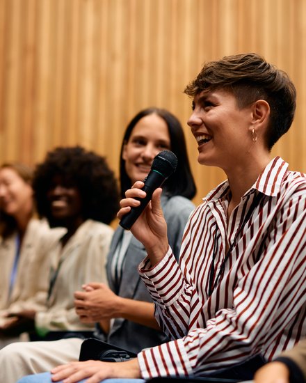 Teilnehmende in einem Vortragssaal sitzen in einer Reihe; im Vordergrund lächelt eine Person im gestreiften Hemd und spricht mit einem Handmikrofon, daneben hören weitere zu.