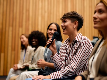 Teilnehmende in einem Vortragssaal sitzen in einer Reihe; im Vordergrund lächelt eine Person im gestreiften Hemd und spricht mit einem Handmikrofon, daneben hören weitere zu.