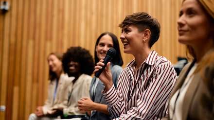 Teilnehmende in einem Vortragssaal sitzen in einer Reihe; im Vordergrund lächelt eine Person im gestreiften Hemd und spricht mit einem Handmikrofon, daneben hören weitere zu.