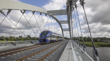 Zug fährt über eine moderne Eisenbahnbrücke mit Stahlbogen und Zugseilen über einen Fluss.