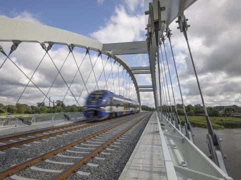 Zug fährt über eine moderne Eisenbahnbrücke mit Stahlbogen und Zugseilen über einen Fluss.