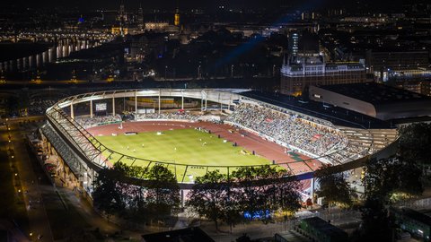 Luftbild des hell erleuchteten Stadions bei Nacht während einer Veranstaltung, umgeben von den Lichtern der nächtlichen Stadt.