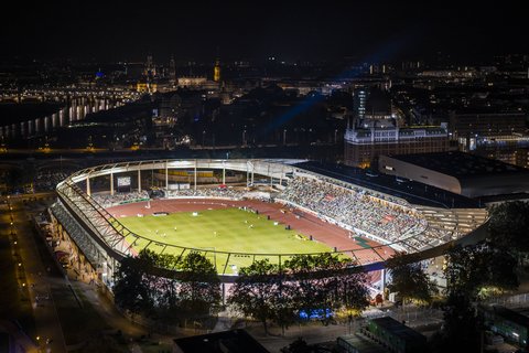 Luftbild des hell erleuchteten Stadions bei Nacht während einer Veranstaltung, umgeben von den Lichtern der nächtlichen Stadt.