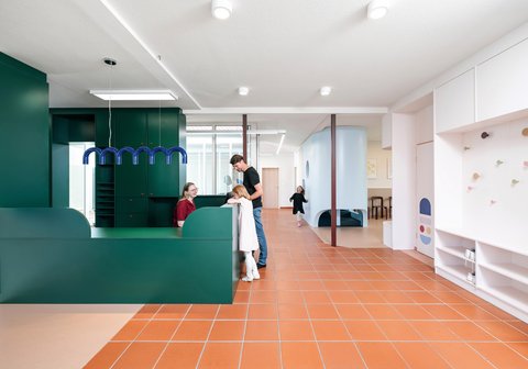 „Ein heller, modern gestalteter Empfangsbereich mit terrakottafarbenem Fliesenboden. Ein Mann und ein Kind stehen an einem markanten, dunkelgrünen Tresen, hinter dem eine Person sitzt.“