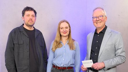 Drei Personen posieren vor grauer Wand mit blauer Beleuchtung; die rechte Person hält eine Karte mit dem Logo der Architektenkammer Nordrhein‑Westfalen.
