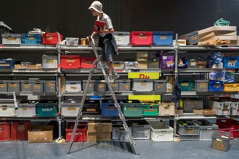 "Person mit weißem Helm steht auf einer großen Metallleiter vor einer hohen Wand aus metallischen Lagerkisten in Rot, Blau, Grün und Grau. Die Kisten sind in mehreren Reihen übereinander gestapelt. Ein dunkler Boden und schwarze Wand im Hintergrund deuten auf einen Lagerbereich hin."