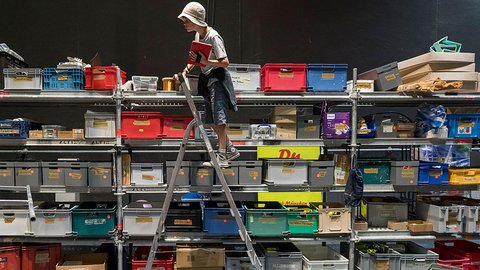 "Person mit weißem Helm steht auf einer großen Metallleiter vor einer hohen Wand aus metallischen Lagerkisten in Rot, Blau, Grün und Grau. Die Kisten sind in mehreren Reihen übereinander gestapelt. Ein dunkler Boden und schwarze Wand im Hintergrund deuten auf einen Lagerbereich hin."