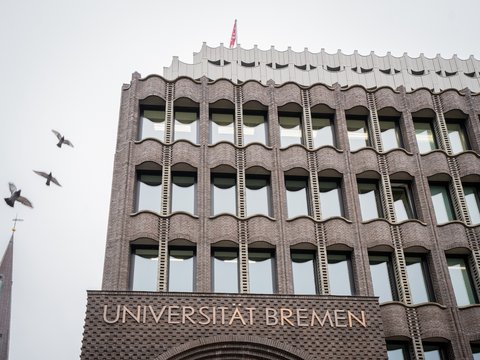Fassade eines mehrstöckigen Backsteingebäudes mit der Aufschrift 'UNIVERSITÄT BREMEN' und mehreren Fenstern, darüber fliegen drei Vögel