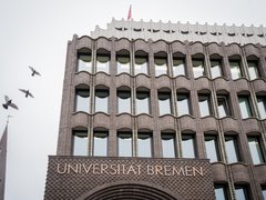 Fassade eines mehrstöckigen Backsteingebäudes mit der Aufschrift 'UNIVERSITÄT BREMEN' und mehreren Fenstern, darüber fliegen drei Vögel