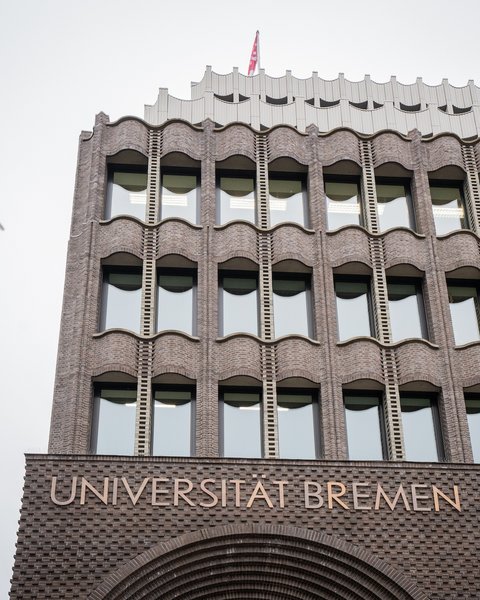 Fassade eines mehrstöckigen Backsteingebäudes mit der Aufschrift 'UNIVERSITÄT BREMEN' und mehreren Fenstern, darüber fliegen drei Vögel