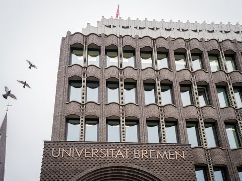 Fassade eines mehrstöckigen Backsteingebäudes mit der Aufschrift 'UNIVERSITÄT BREMEN' und mehreren Fenstern, darüber fliegen drei Vögel