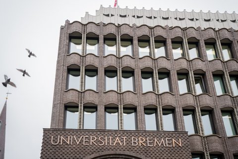 Fassade eines mehrstöckigen Backsteingebäudes mit der Aufschrift 'UNIVERSITÄT BREMEN' und mehreren Fenstern, darüber fliegen drei Vögel