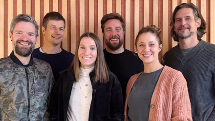 Sechs Personen stehen nebeneinander vor einer vertikalen Holzlamellenwand und lächeln in die Kamera.