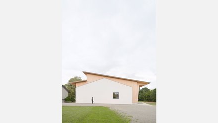 Moderne Gemeindehalle mit weißer Fassade und Holzverkleidung, davor eine Person auf einem Gehweg, umgeben von Rasen und Bäumen
