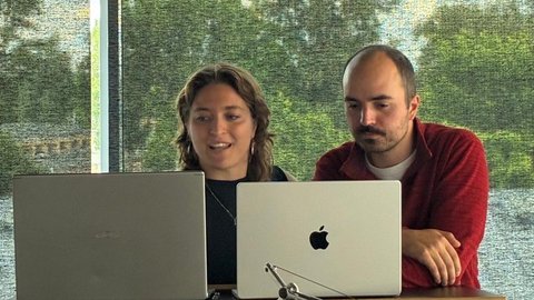 Zwei Personen arbeiten zusammen an einem Schreibtisch vor einem großen Fenster. Eine Frau mit braunem, welligem Haar sitzt links und lächelt, sie trägt ein schwarzes Oberteil. Ein Mann mit rötlichem Bart sitzt rechts in einem dunkelroten Hemd. Zwischen ihnen stehen zwei aufgeklappte Laptops auf einem Holztisch. Im Hintergrund ist eine grüne Landschaft mit Bäumen durch das Fenster zu sehen. Auf dem Tisch liegen weitere Materialien und ein Glas Wasser.