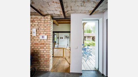 Moderner Innenraum eines renovierten Altbaus mit Backsteinmauer links, dunklen Holzbalken an der Decke und weißen Wänden. Ein Holzgitter trennt einen unteren Bereich ab. Durch eine große Glastür rechts ist eine Terrasse mit blauen Gartenstühlen und grüner Vegetation sichtbar. An der Backsteinwand hängen zwei kleine Bilder. Der Boden ist mit hellen Holzdielen und grauem Betonstein belegt.