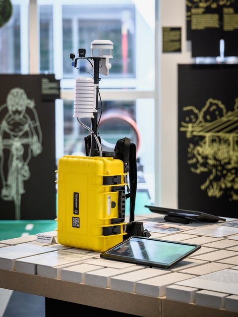 "Gelber wetterfester Kunststoffkoffer auf einer Ausstellungsfläche. Darauf montiert ein schwarzes Gestell mit weißem zylindrischem Messinstrument zur Klimadatenerfassung. Ein digitales Tablet liegt auf der Fläche darunter. Im Hintergrund schwarze Ausstellungswand und verschwommene Museumsräume."
