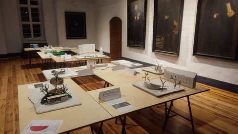 "Ein Blick in einen Ausstellungsraum mit Holzboden und weißen Wänden. Auf mehreren Tischen, die über den Raum verteilt sind, stehen filigrane Architekturmodelle, die meisten davon weiß und minimalistisch. An der rechten Wand hängen drei große, dunkle Porträtgemälde in goldenen Rahmen, die historische Persönlichkeiten zeigen."