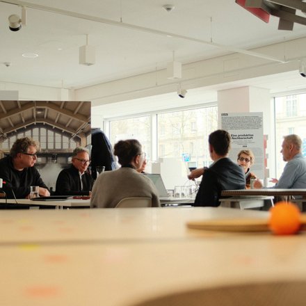 Mehrere Personen sitzen in einem hellen Raum an einem Tisch und diskutieren. Im Hintergrund sind Ausstellungswände und große Fenster zu sehen.