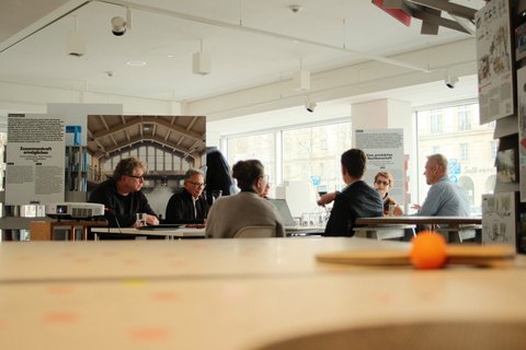 Mehrere Personen sitzen in einem hellen Raum an einem Tisch und diskutieren. Im Hintergrund sind Ausstellungswände und große Fenster zu sehen.