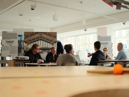 Mehrere Personen sitzen in einem hellen Raum an einem Tisch und diskutieren. Im Hintergrund sind Ausstellungswände und große Fenster zu sehen.
